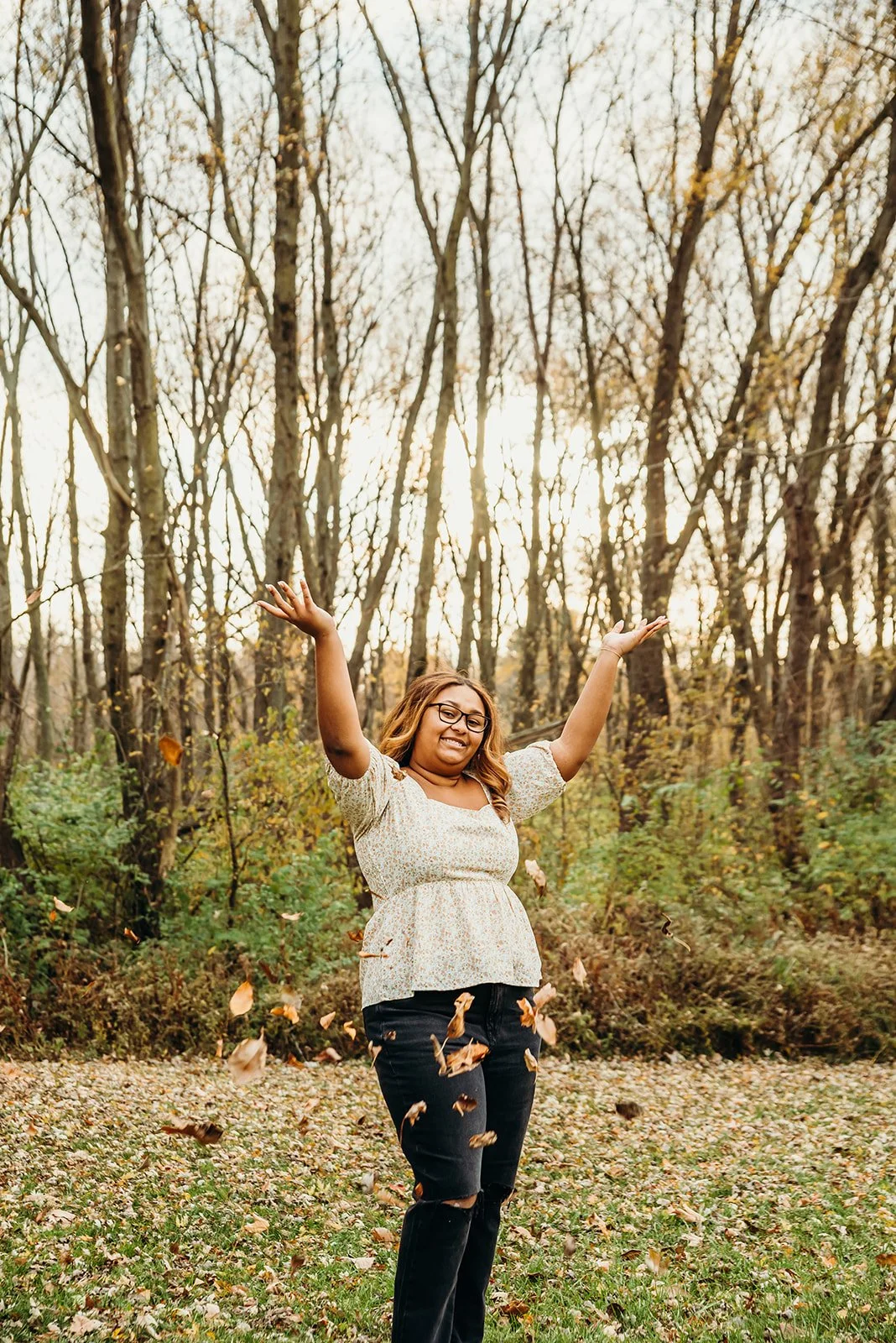 A woman with glasses and long hair smiling with arms raised, standing in a park with trees and fallen leaves during autumn, with the sun setting in the background.
