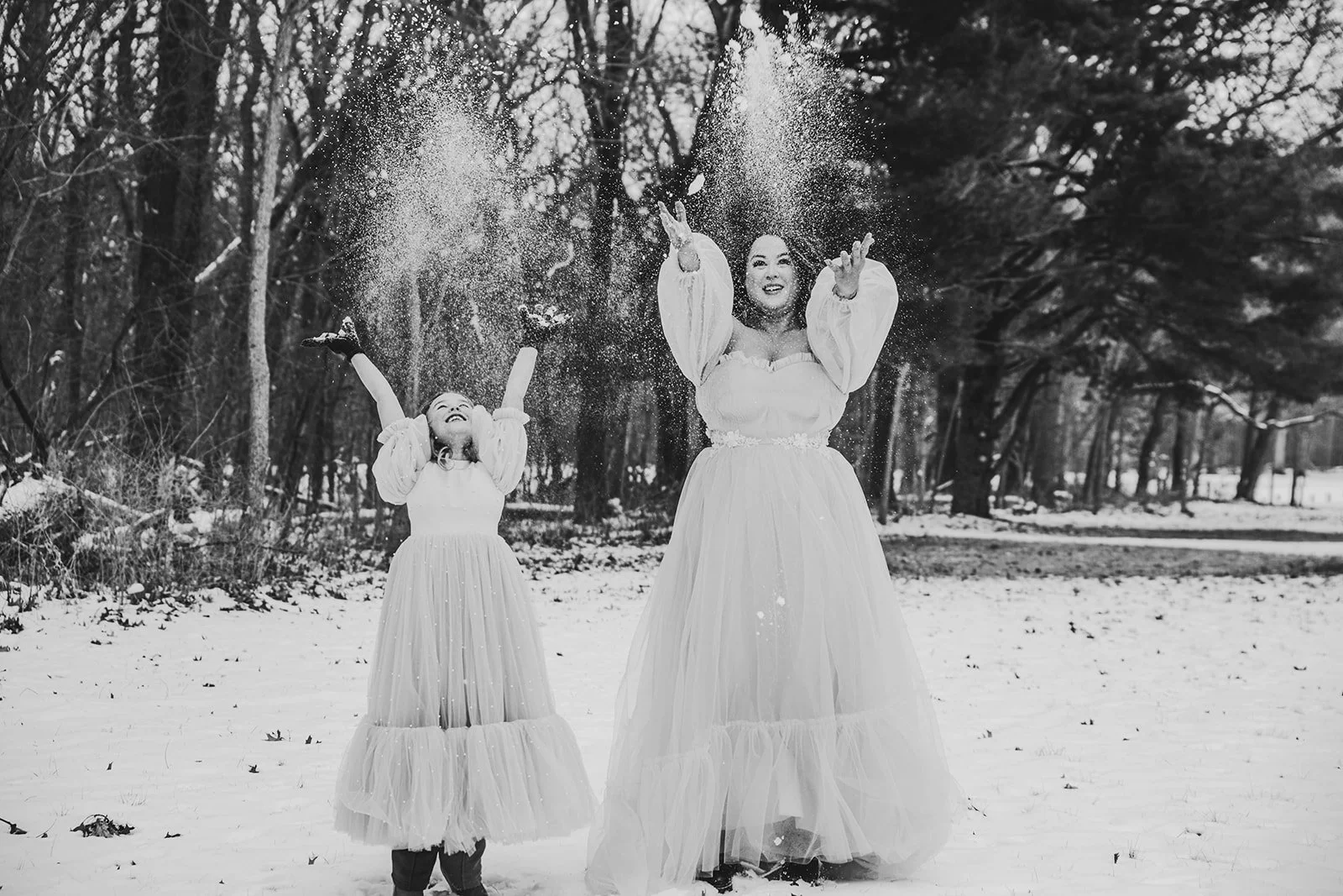 Two women wearing long, flowing dresses standing outdoors in a snow-covered park with bare trees in the background. They are throwing snow into the air, and both are smiling.