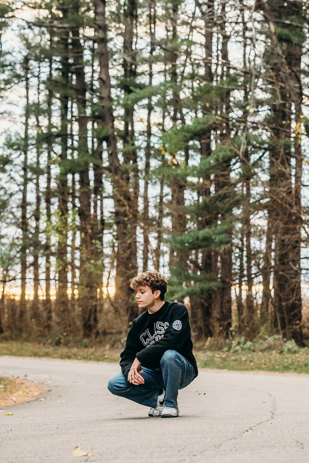 A young person with curly hair squatting on a paved road in a forest during sunset, wearing a black sweatshirt, blue jeans, and sneakers.