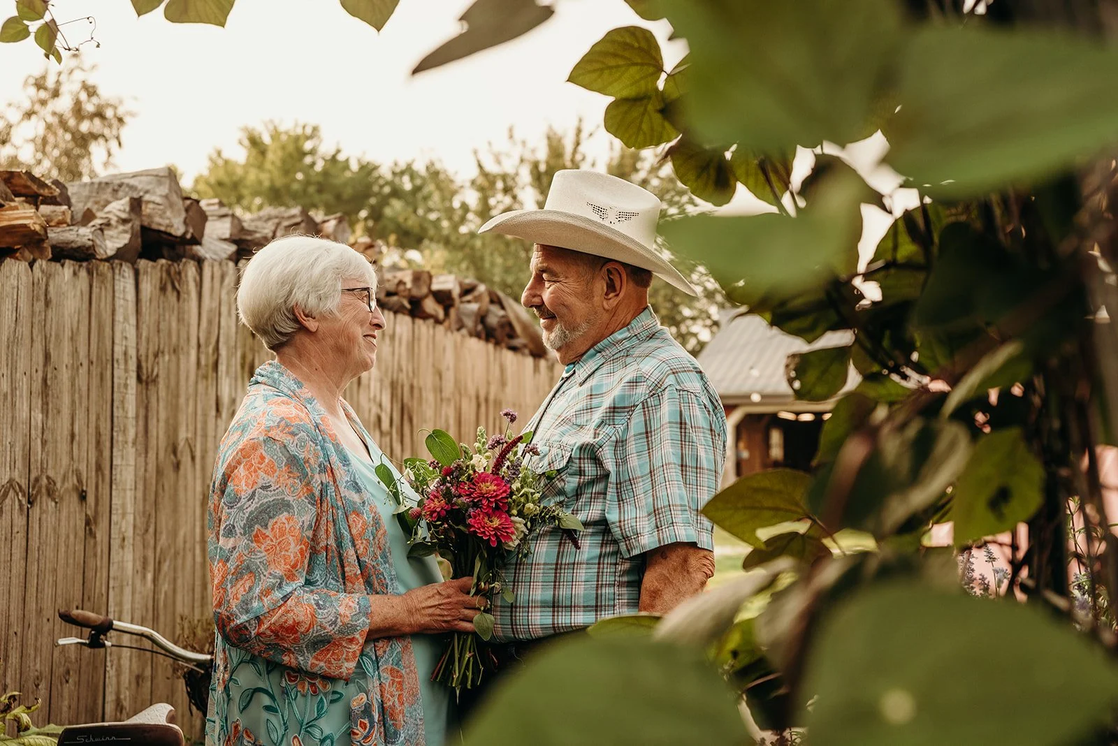 An elderly couple stands outdoors, exchanging smiles. The woman holds a bouquet of pink and purple flowers, and they are framed by green leaves and a wooden fence behind them.