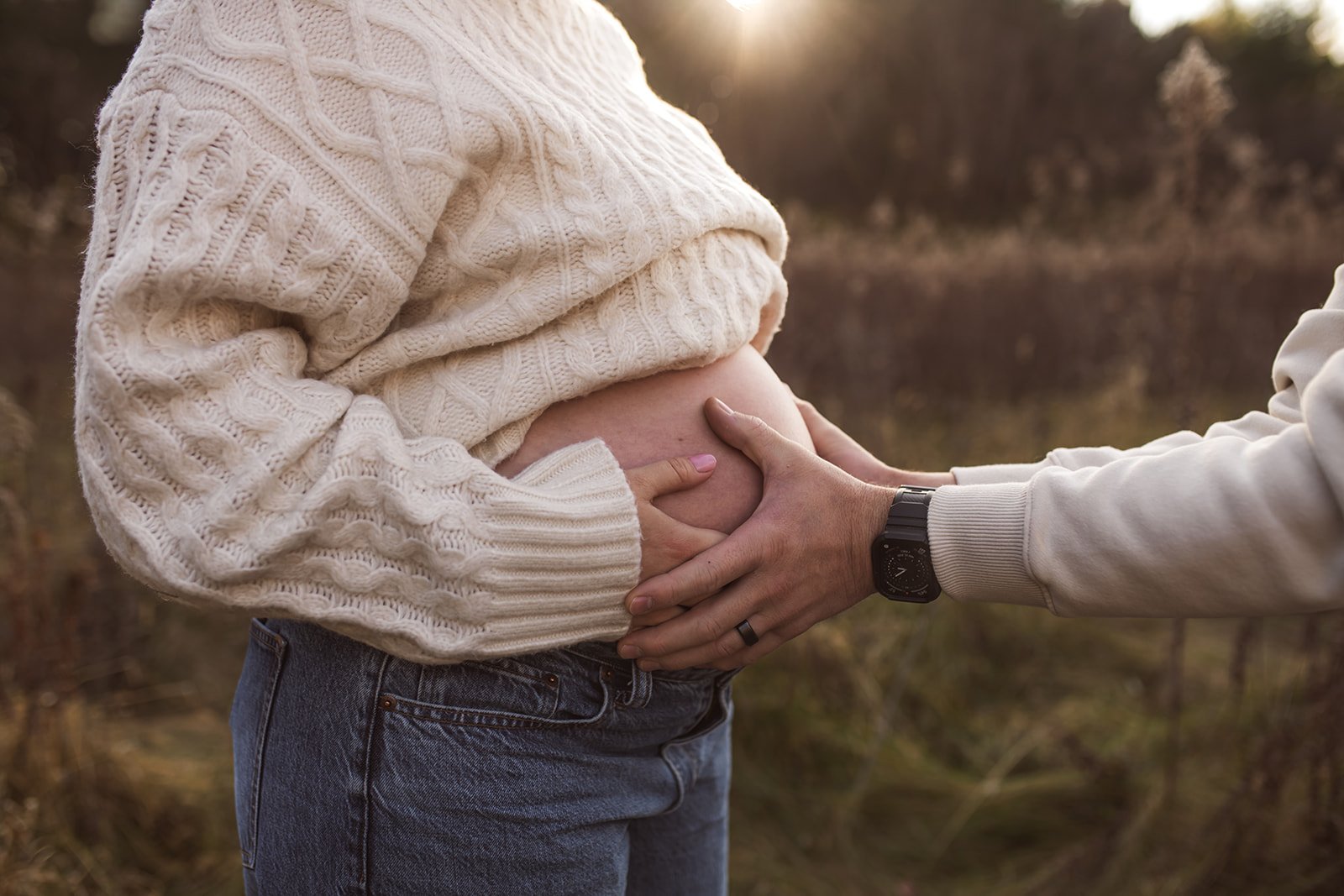 A pregnant woman in a cream cable-knit sweater holding her belly, with another person holding her belly from the side, outdoors during sunset.