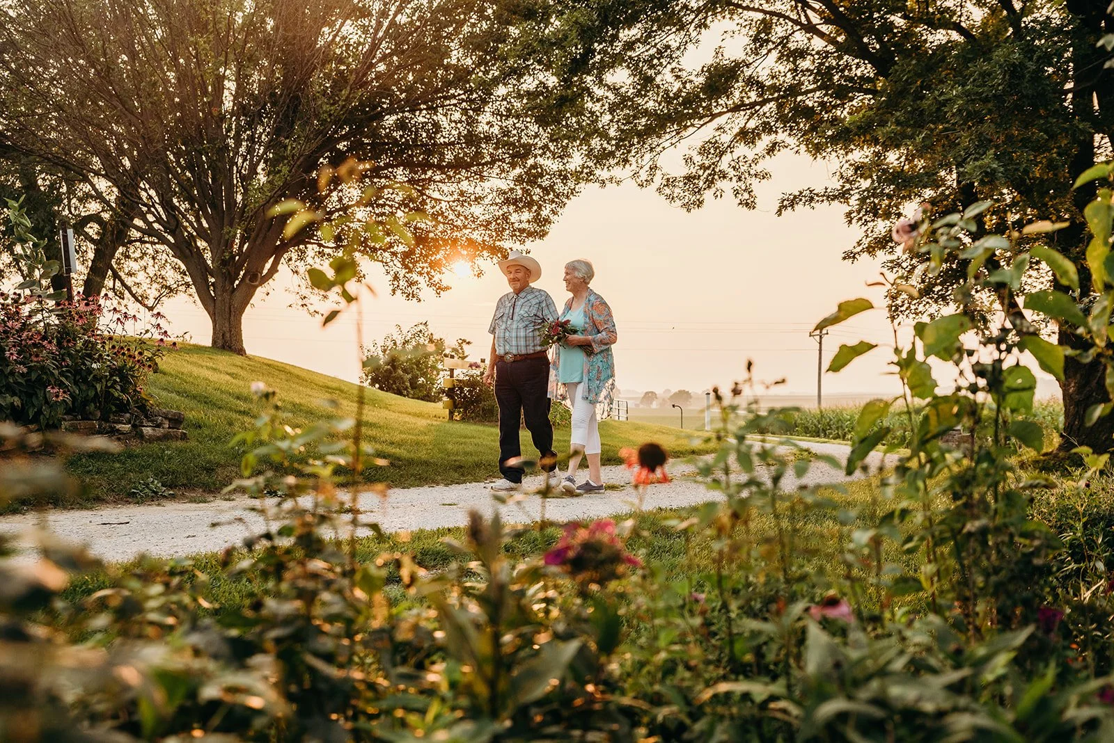 An elderly couple walking on a garden path during sunset, surrounded by trees and flowering plants, with a rural landscape in the background.
