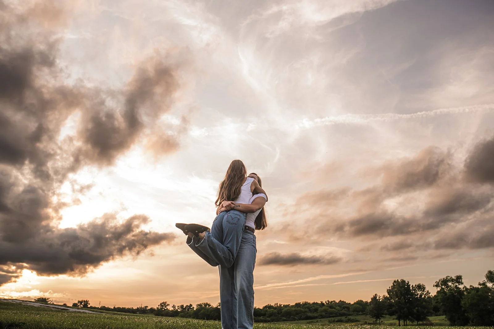 Two women hugging outdoors at sunset with cloudy sky and trees in the background.