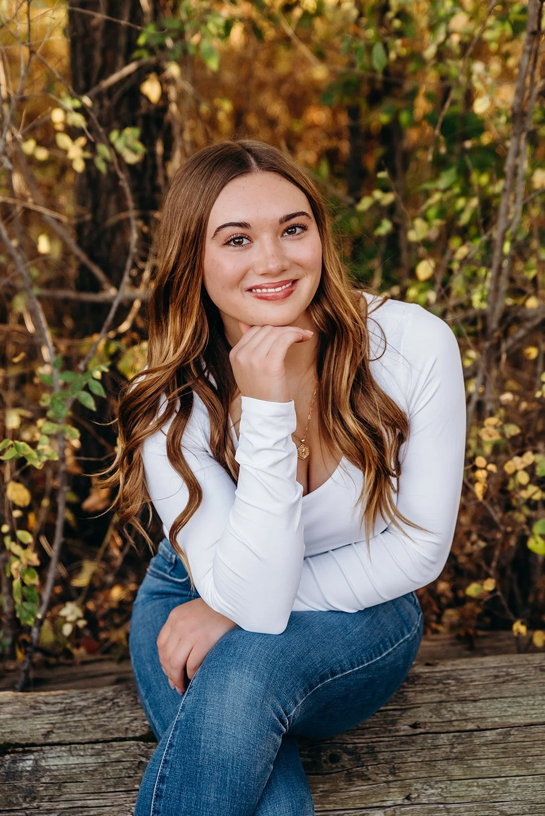 A young woman with long wavy brown hair, wearing a white long-sleeve shirt and blue jeans, sitting on a wooden log outdoors with autumn leaves and foliage in the background, smiling and resting her chin on her hand.
