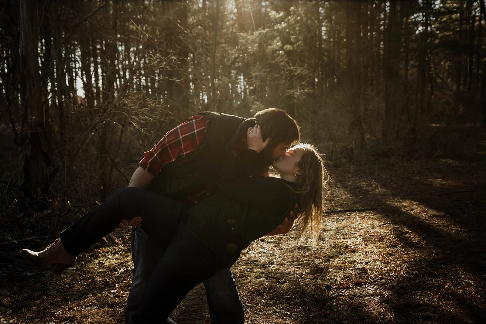 A couple sharing a kiss in a forest during sunset, with the man holding the woman in his arms.