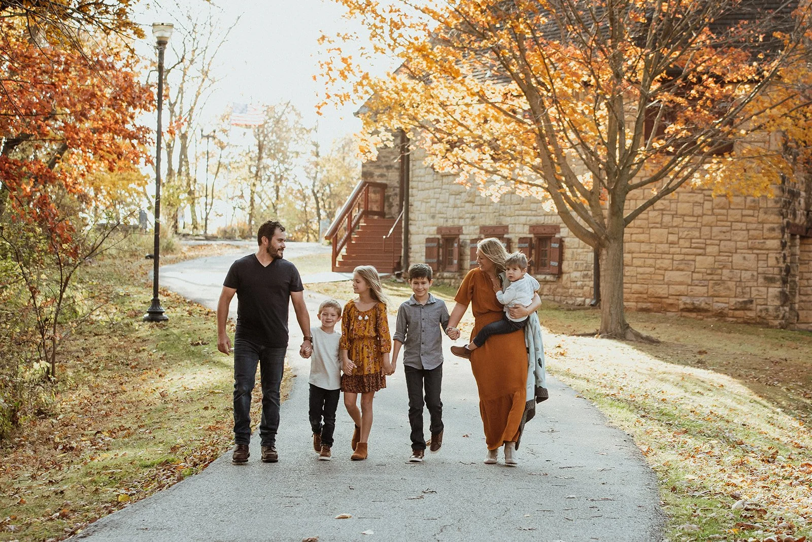 A family of six walking outdoors on a fall day, holding hands, beside a house with autumn trees.