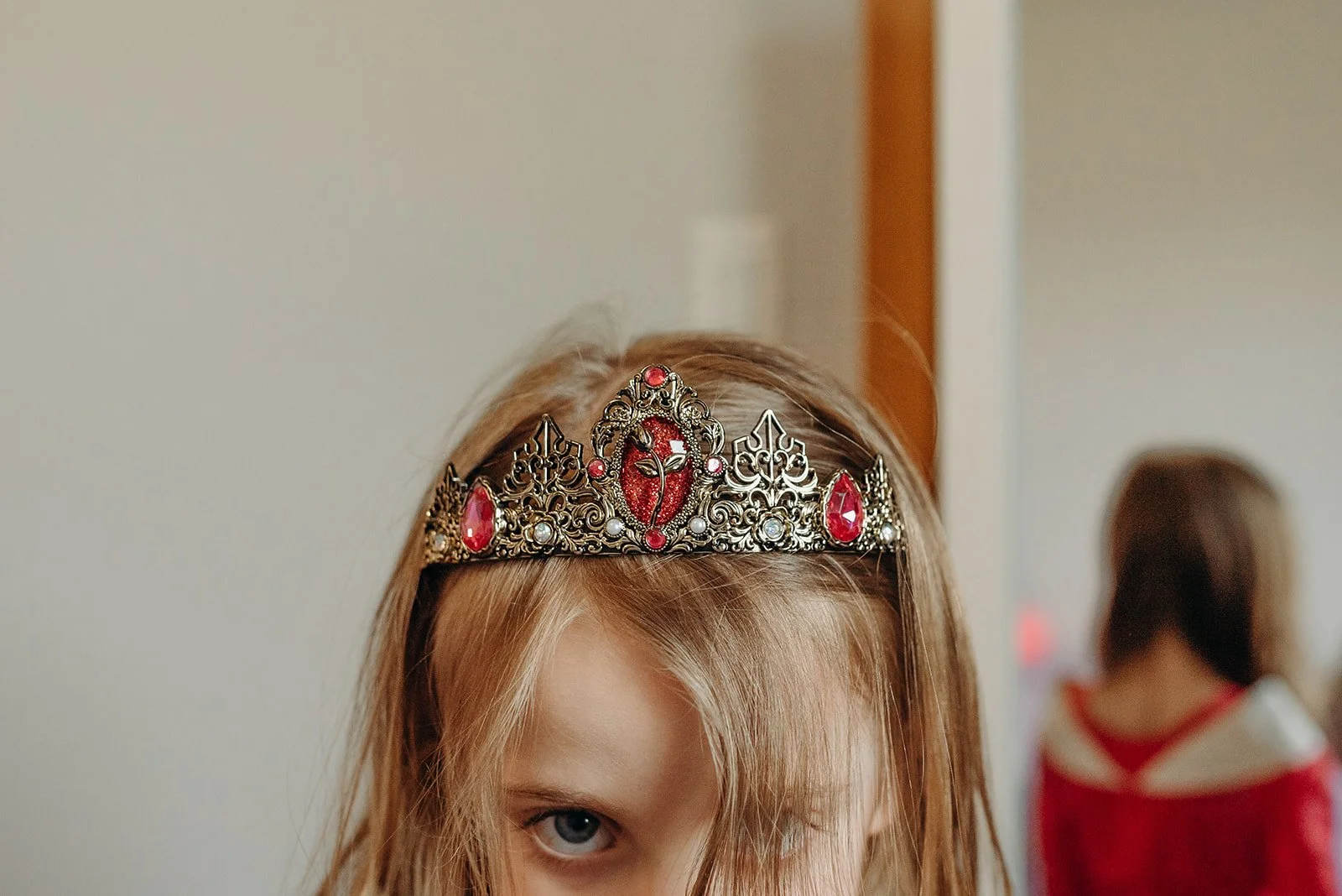 Child wearing a decorative crown with red gemstones, in front of a mirror.