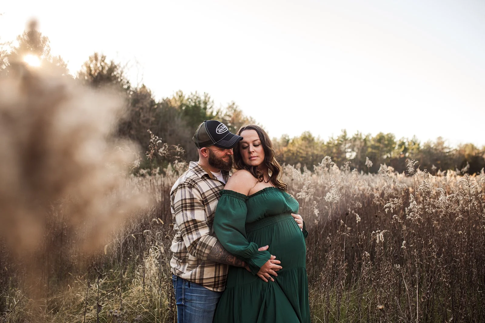 A pregnant woman in a green dress standing in a field with a man wearing a plaid shirt and baseball cap. They are embracing, with the man's hands on her belly, and she has her eyes closed, looking contemplative. The scene is set in late afternoon or 
