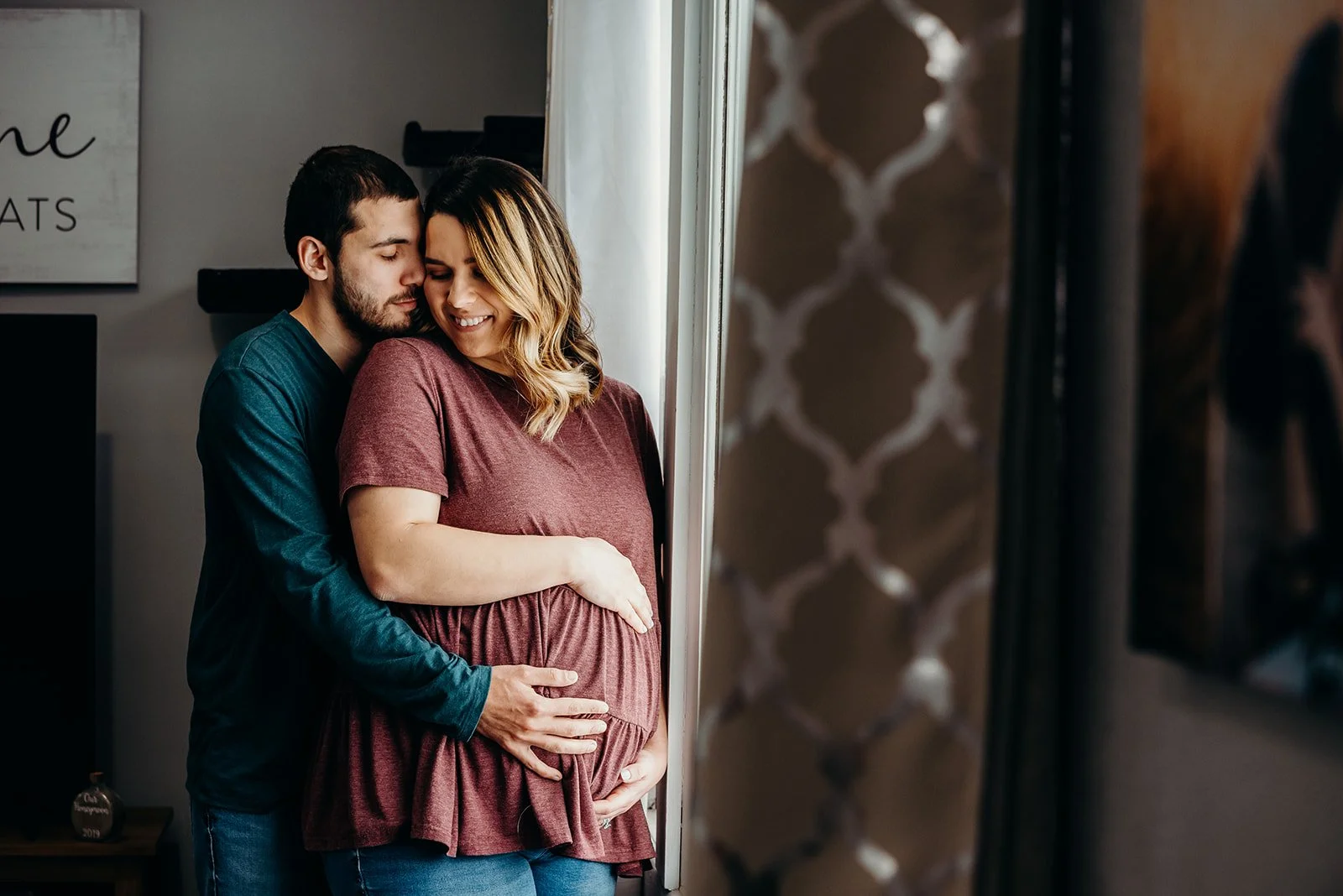 A couple standing close by a window, with the man hugging the pregnant woman from behind. They are both smiling and looking down at her baby bump.
