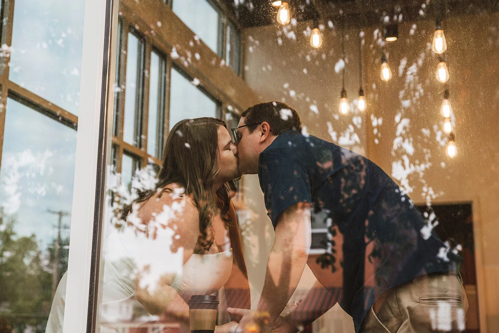 A couple kissed through a glass window, with reflections of trees and lights visible in the glass.