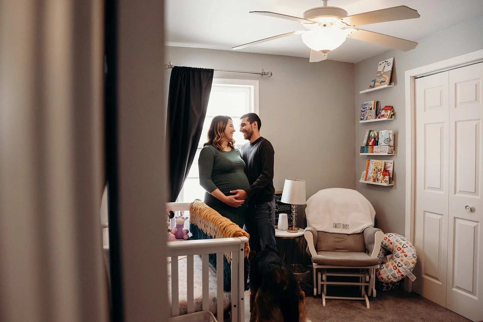 A couple stands in a nursery, smiling and looking into each other's eyes with their hands on a pregnant woman's belly, near a crib and a rocking chair, with a bookshelf and a ceiling fan in the background.