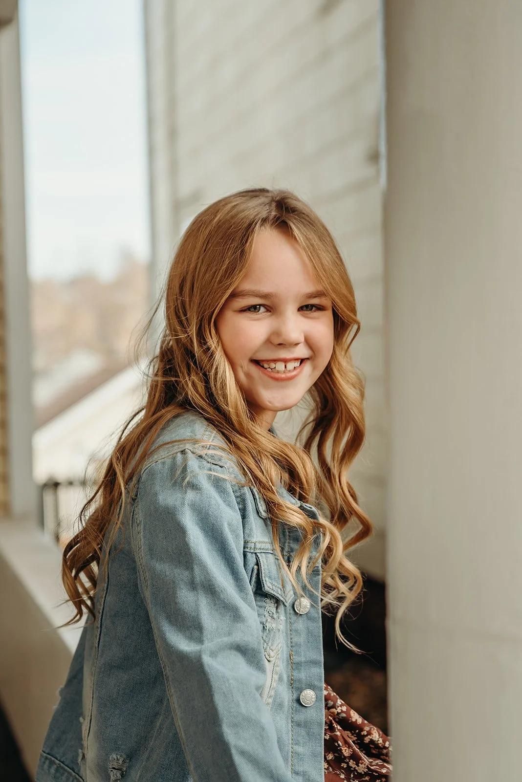 Smiling young girl with long, wavy, light brown hair wearing a denim jacket, standing near a window with a blurred outdoor background.
