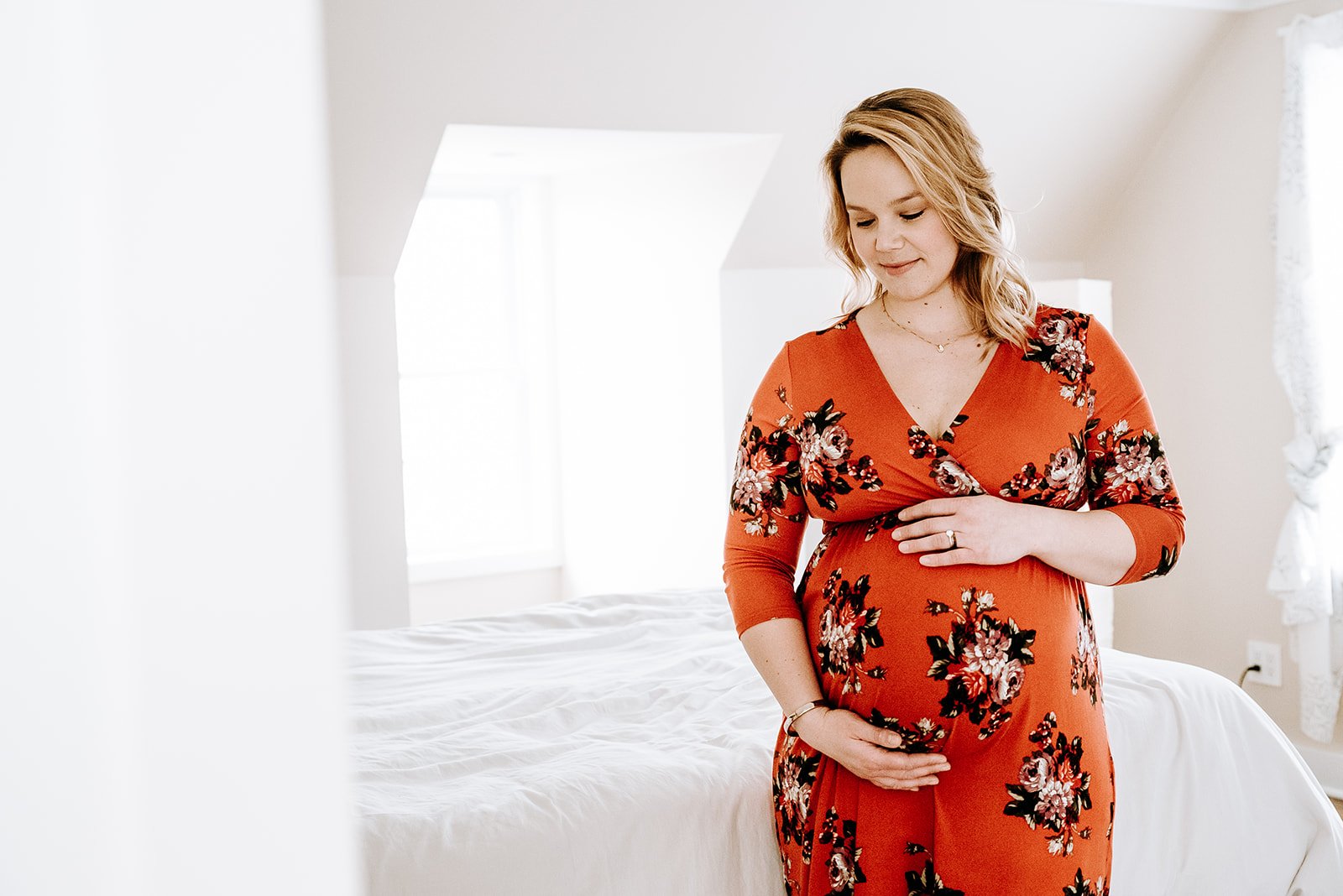 Pregnant woman in a floral red dress gently holding her belly inside a bright bedroom.