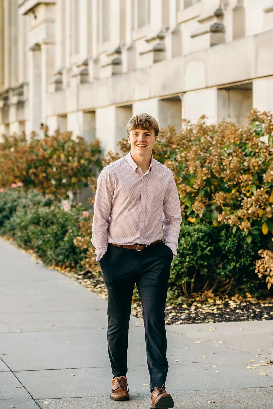 A young man with light brown hair wearing a light pink dress shirt, dark trousers, and brown shoes walking outdoors on a paved sidewalk with bushes and a building in the background.