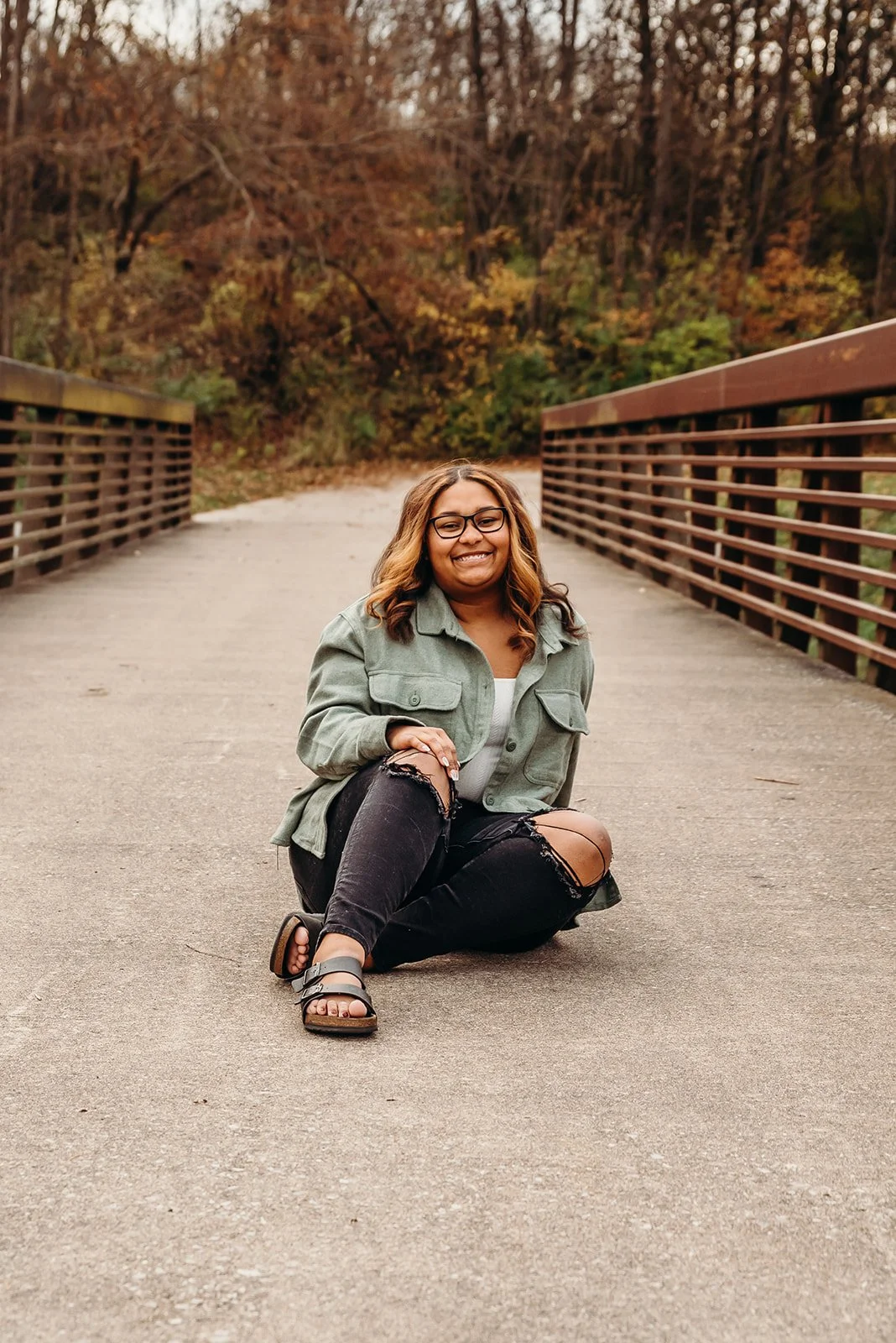 A woman sitting on a park bridge with fall-colored trees in the background, smiling at the camera.