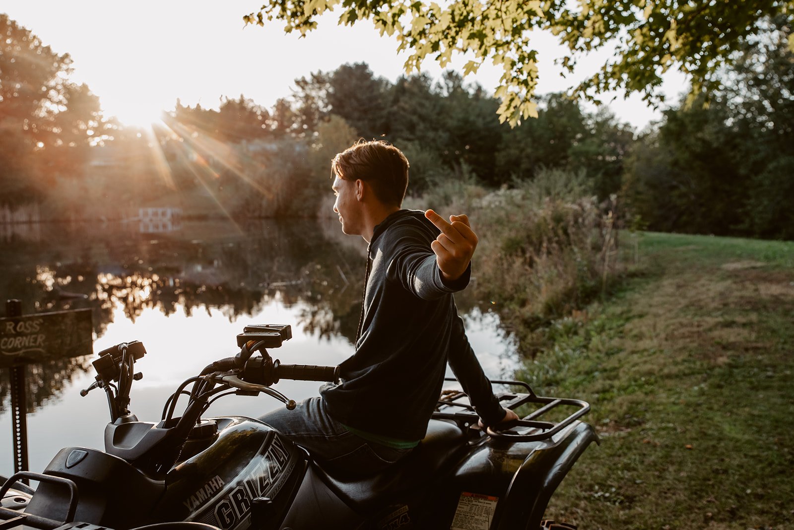 A young man sitting on a Yamaha all-terrain vehicle (ATV) near a lake during sunset, making a gesture with his middle finger.