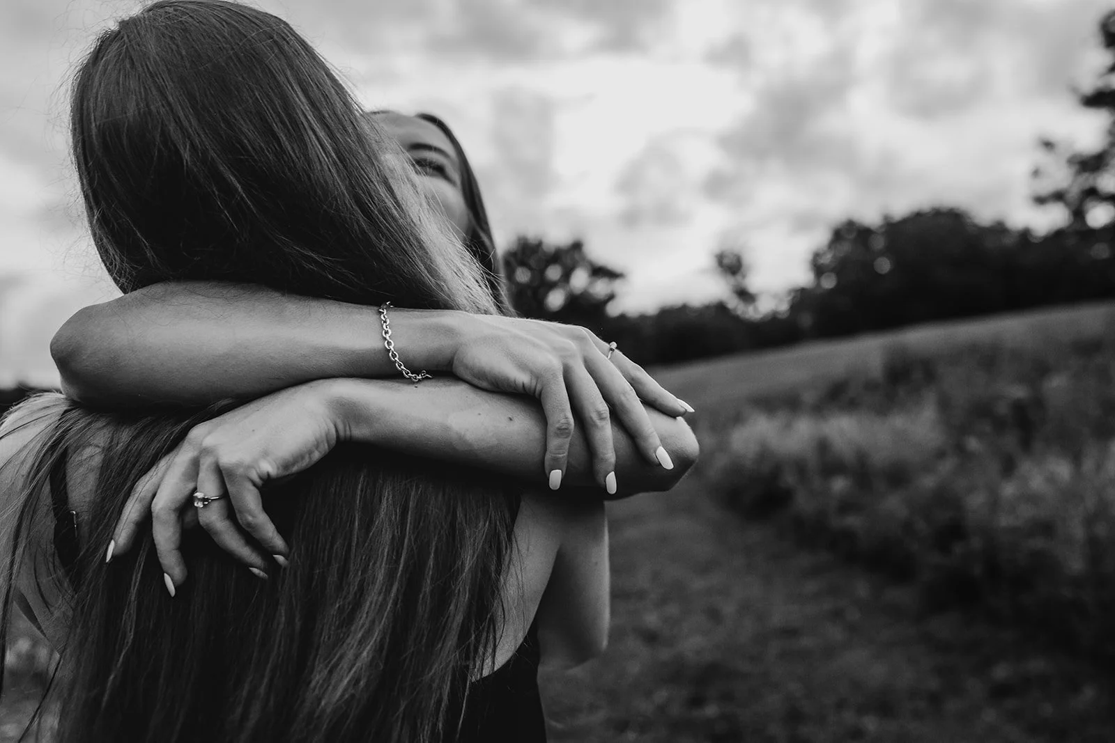 Two women hugging outdoors, black and white photo with trees and sky in background.