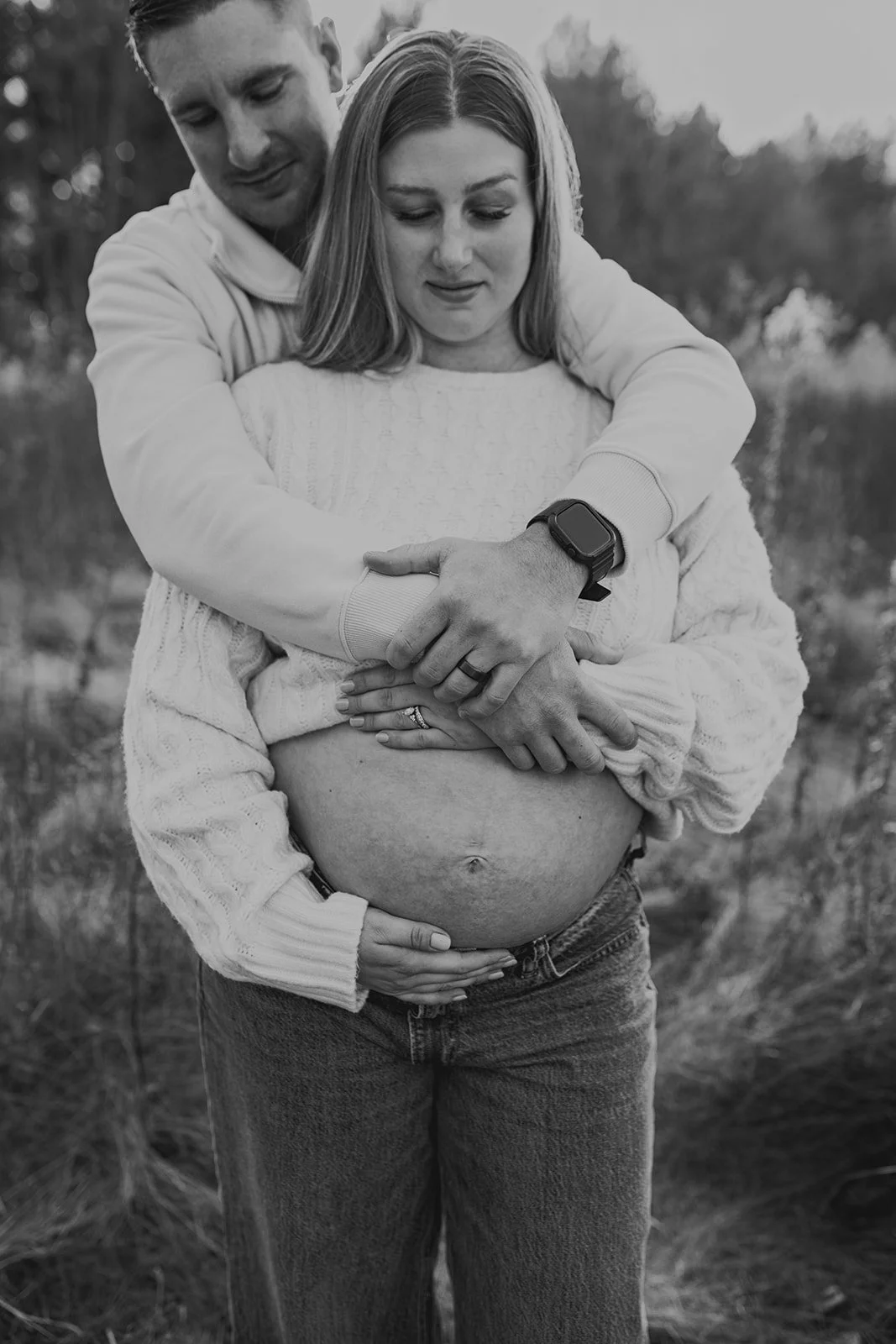 A pregnant woman with a man embracing her from behind, outdoors. The man has his arms around her, with one hand on her pregnant belly.