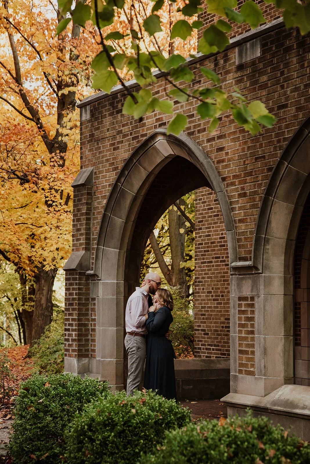 A couple sharing an intimate moment under a brick archway surrounded by autumn trees and greenery.