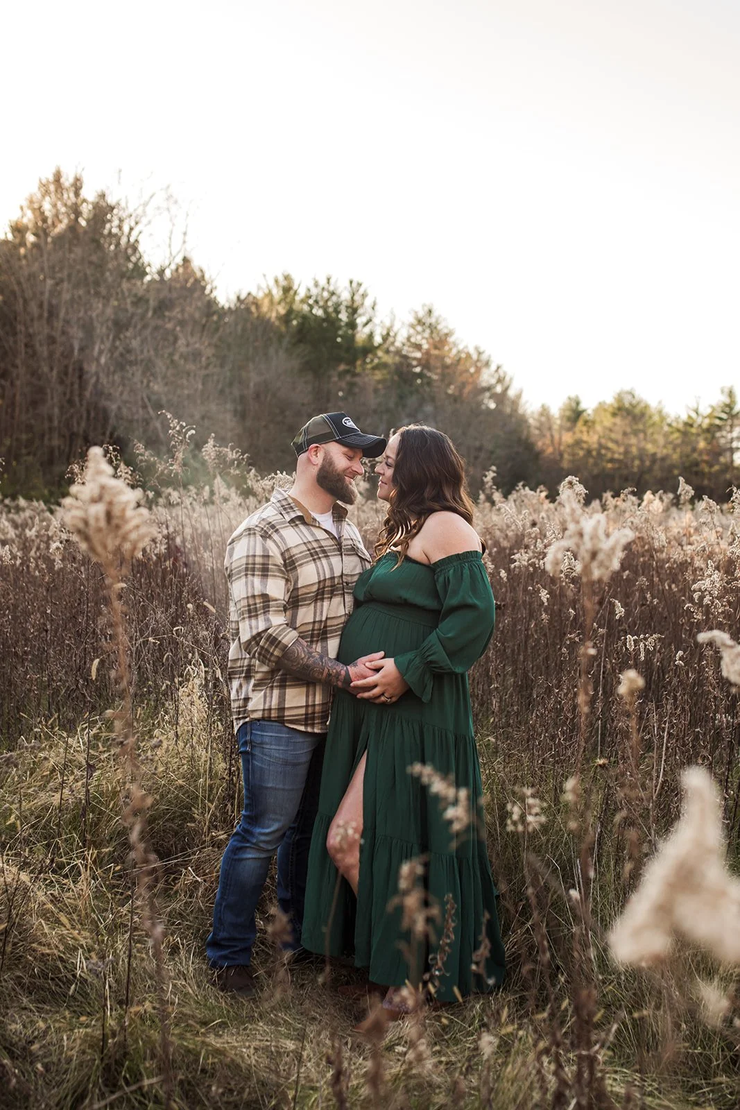 A couple stands in a field of tall, dry grass and wildflowers, facing each other and smiling, with the woman's hands resting on her pregnant belly. The man wears a plaid shirt and a baseball cap, and the woman wears an off-the-shoulder green dress wi