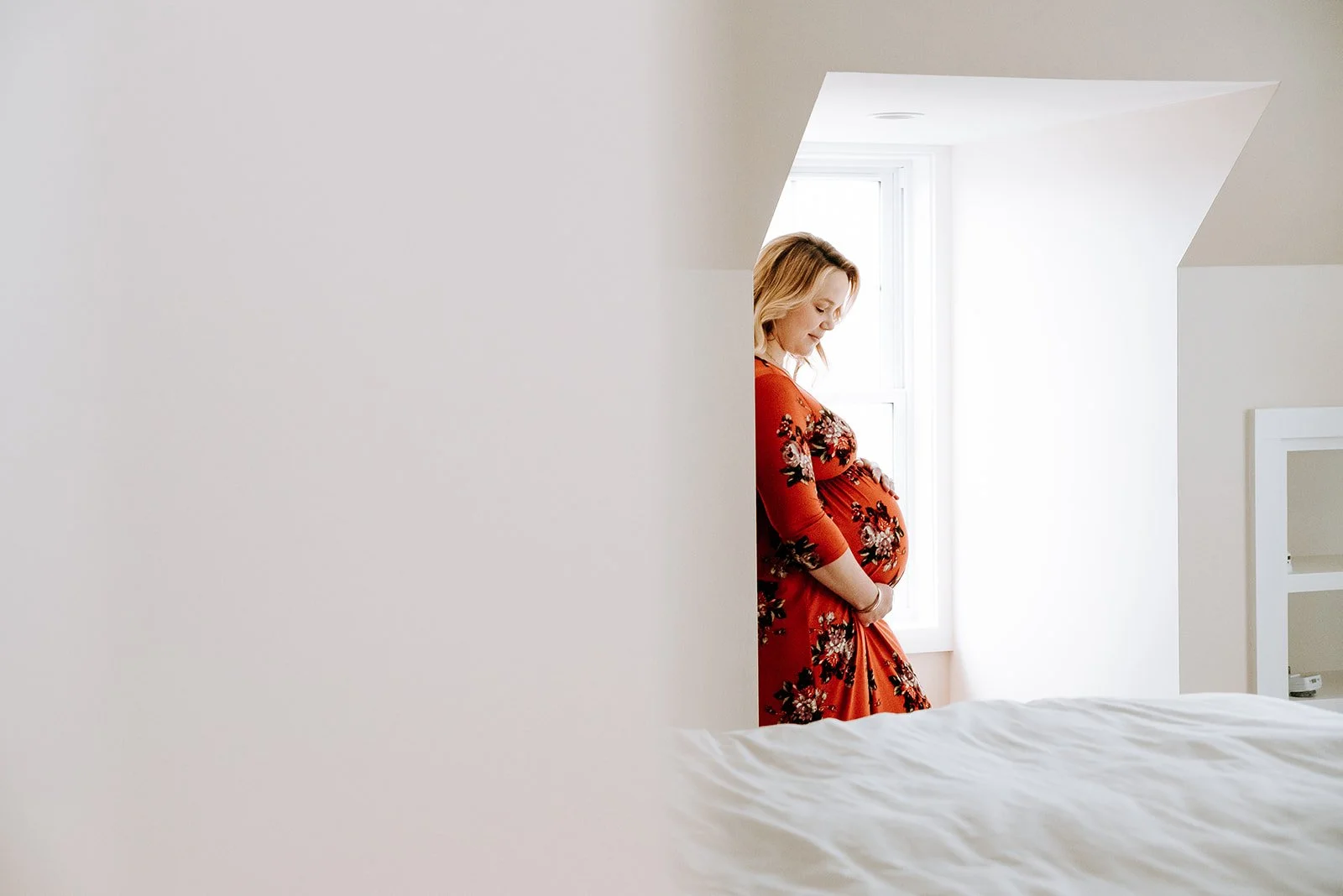 Pregnant woman standing by a window in a brightly lit room, looking down at her belly, wearing a red dress with floral patterns.
