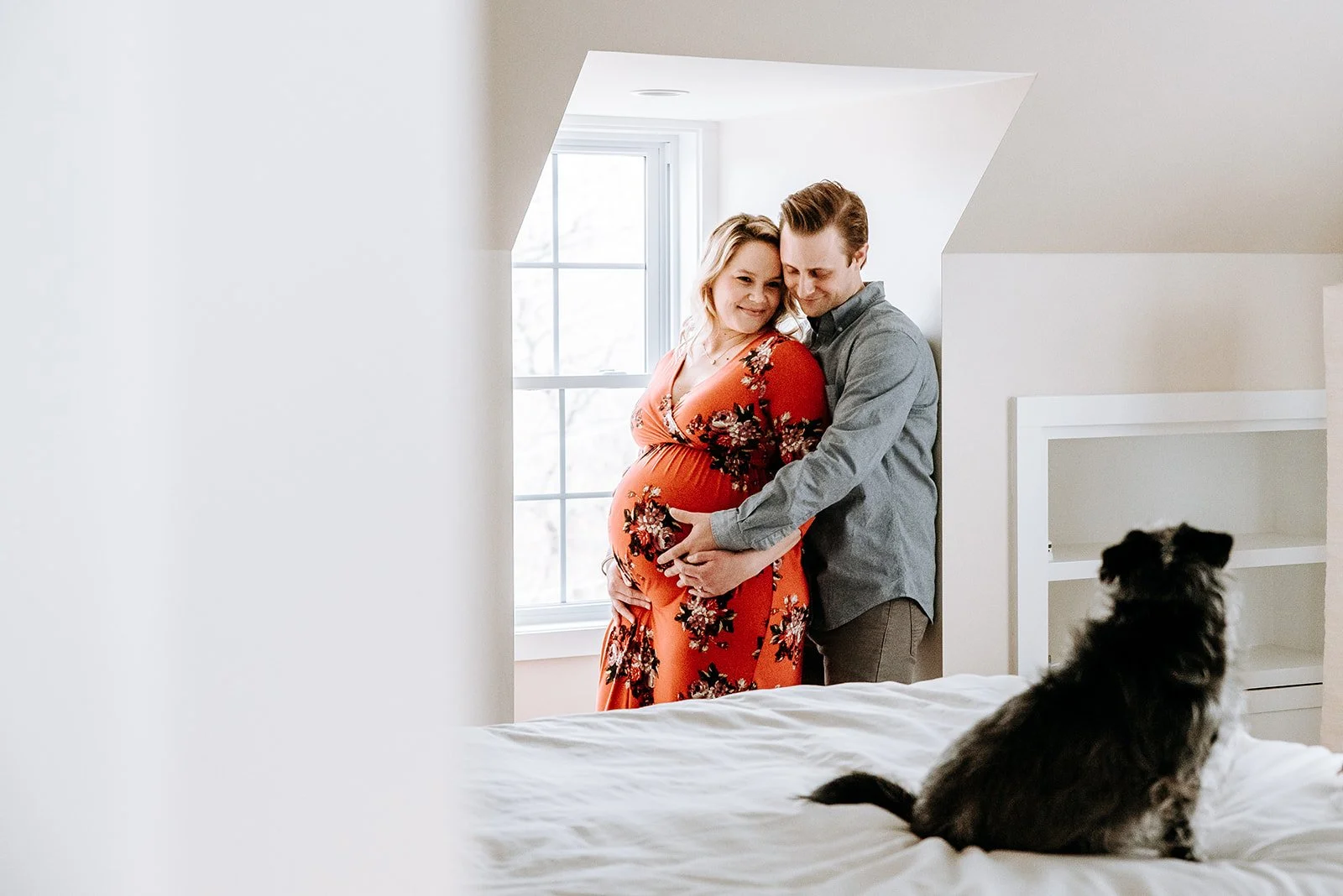 A pregnant woman in a floral orange dress and a man stand together in a bright bedroom, holding her belly. They smile and look down at her belly as a dog sits on the bed, facing away from the camera, with a window in the background.