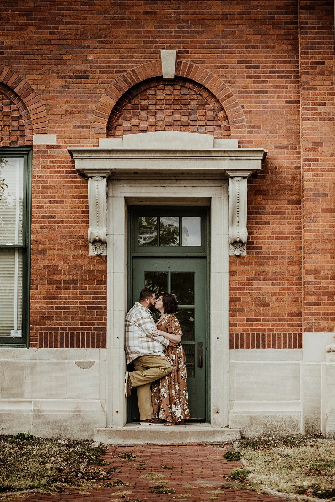 A couple sharing a kiss on a small porch in front of a green door, with a brick building facade behind them.
