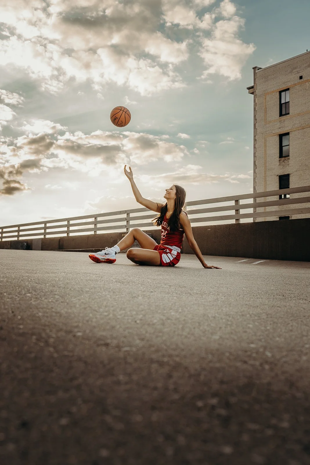 A female basketball player sitting on the ground in a red and white uniform, tossing a basketball into the air on an outdoor parking lot with a building and cloudy sky in the background.