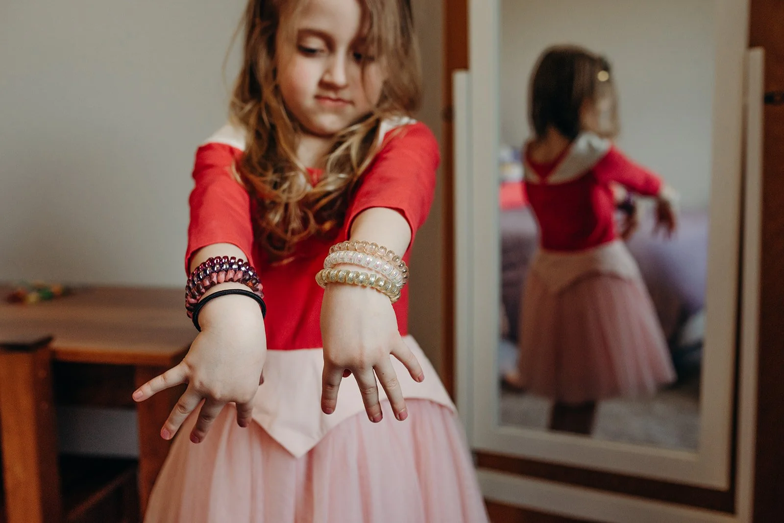 A young girl in a red top with a pink skirt showing her colorful bracelets in front of a mirror, creating a reflection of herself.