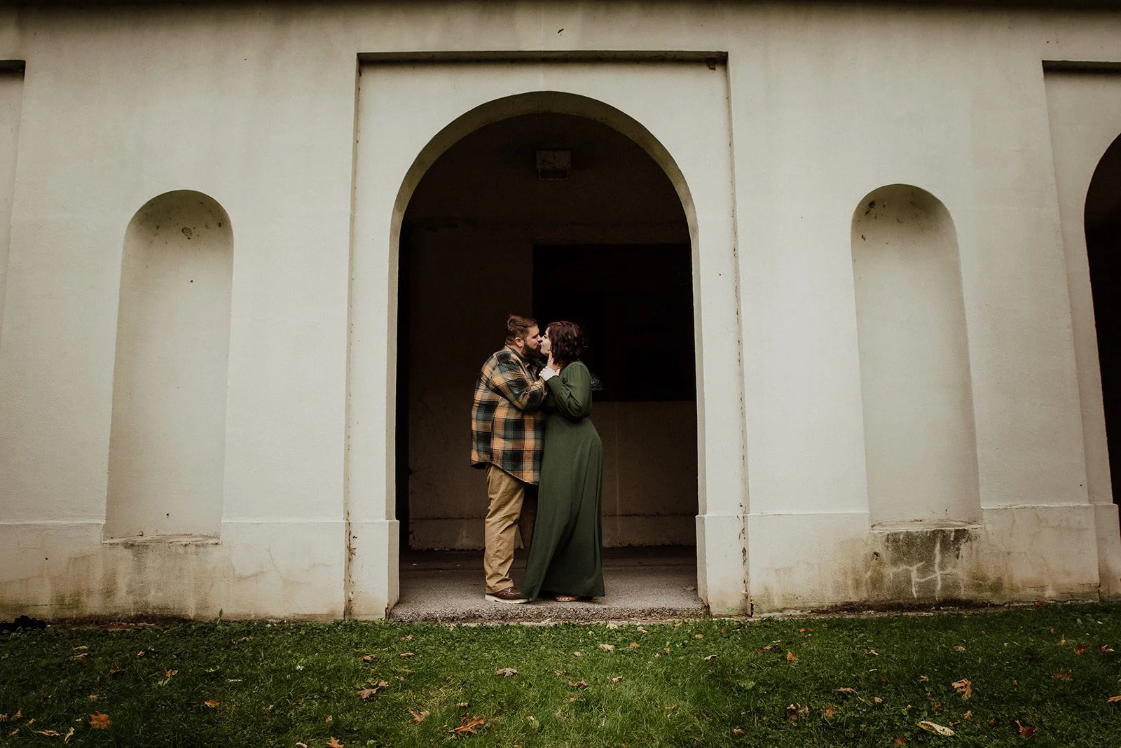 A couple stands close together, about to kiss, inside an arched doorway of a white building with grass and fallen leaves outside.