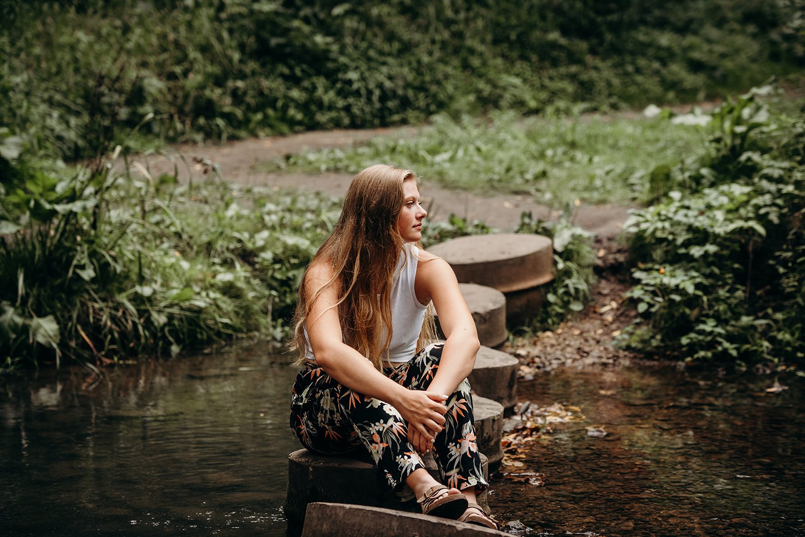 A woman with long, wavy hair sitting on a rock by a small stream in a lush, green outdoor setting.