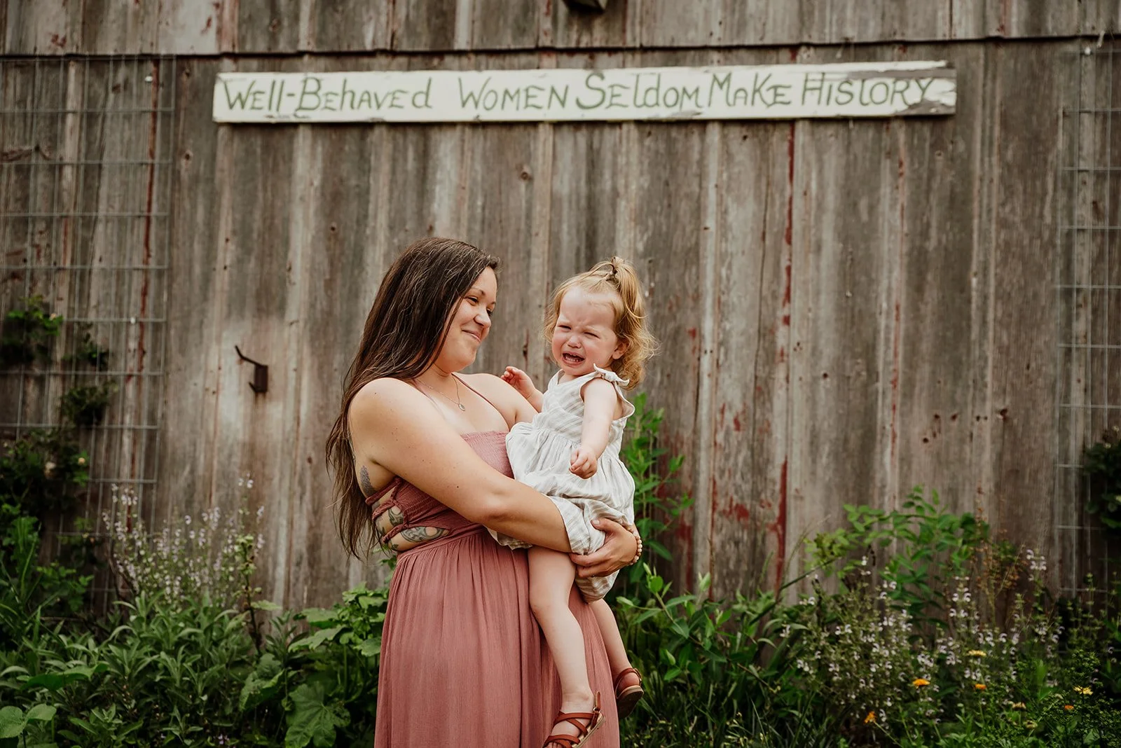 A woman holding a crying young girl in front of a wooden fence with a sign that reads 'Well-Behaved Women Seldom Make History'.