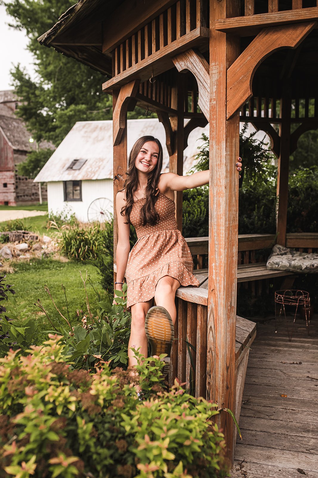 Smiling young woman in a brown polka dot dress sitting on a wooden porch railing surrounded by greenery, with rustic buildings in the background.