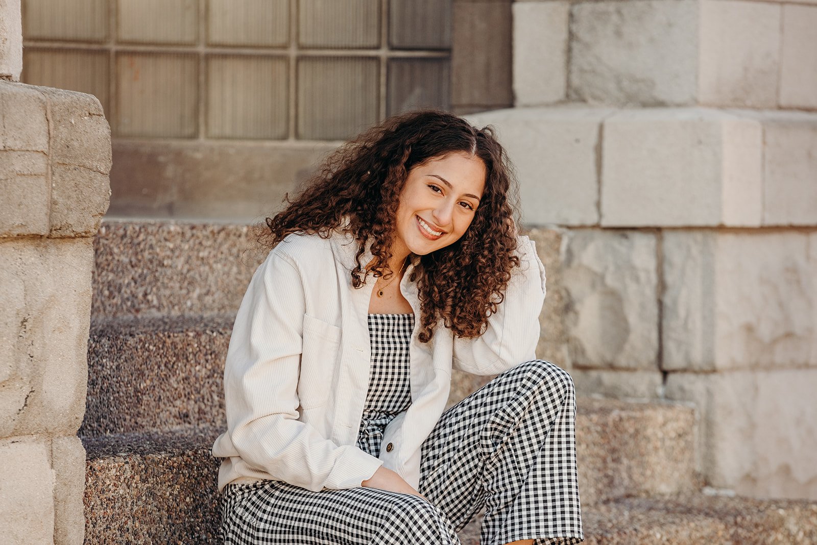 Young woman sitting on stone steps outdoors, smiling, wearing a white jacket, black and white checkered top, and matching checkered pants.