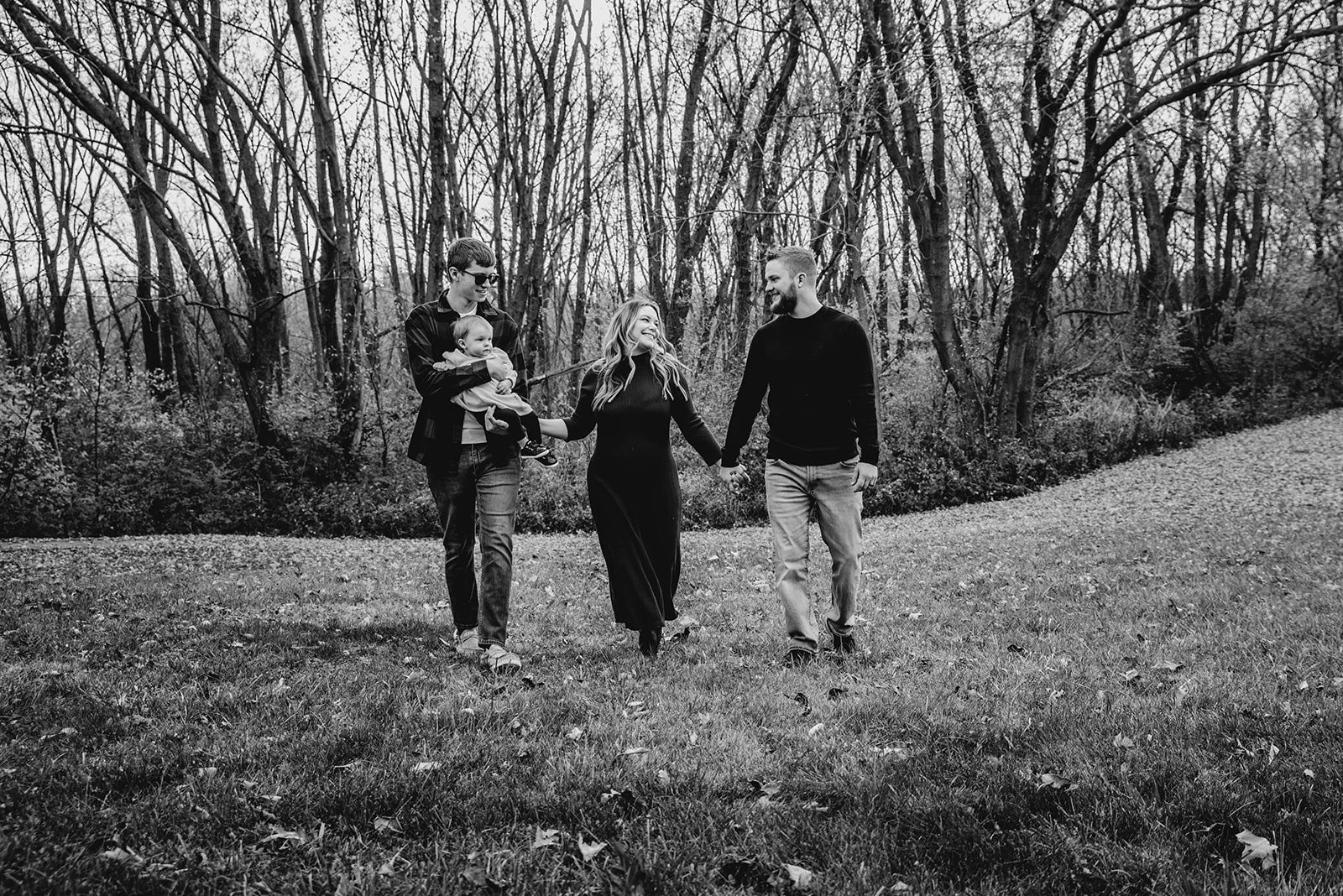 A family of four walking outdoors in a park with leafless trees in the background, holding hands and smiling at each other.