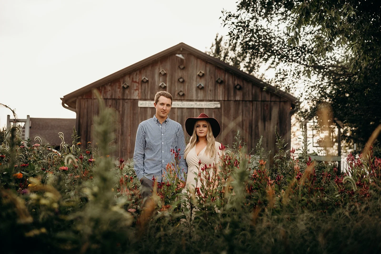A man and woman standing together amidst colorful flowers in front of a rustic wooden barn wearing casual and stylish clothing.