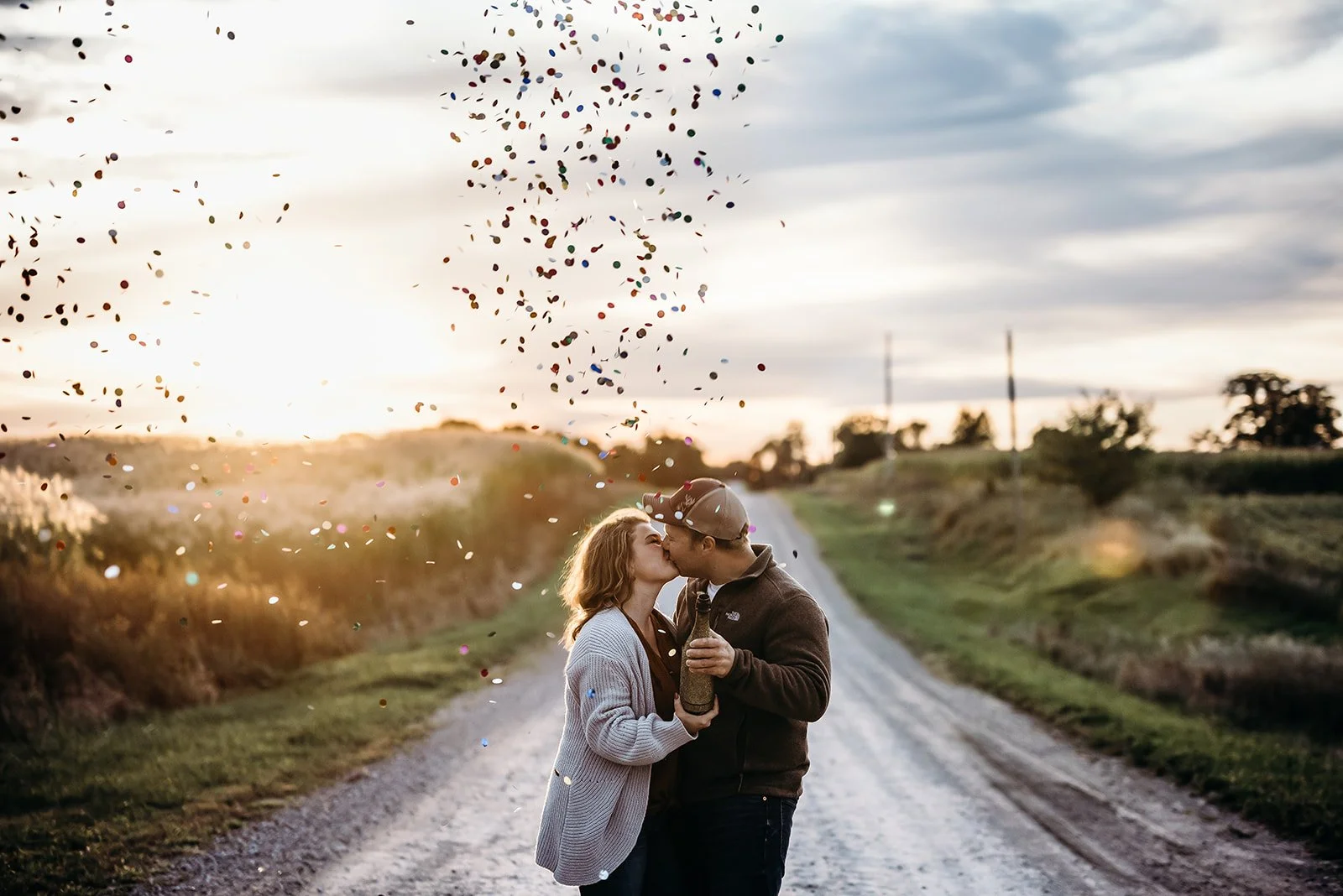 A couple kissing on a dirt road during sunset, celebrating with confetti falling around them.