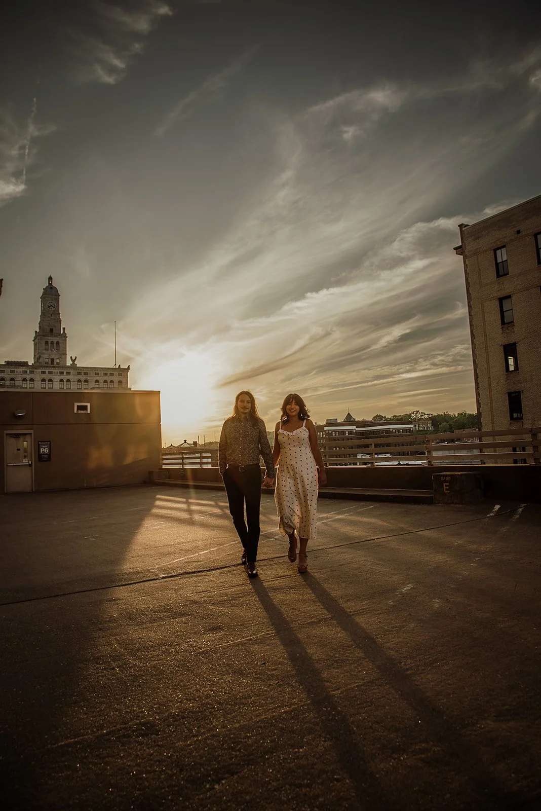Two women walk together on a rooftop parking lot during sunset, with long shadows and a city skyline in the background.