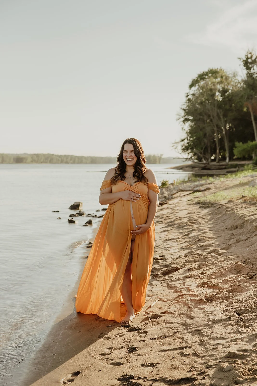 Pregnant woman standing on a sandy beach at sunset, smiling, wearing an orange dress, holding her pregnant belly.