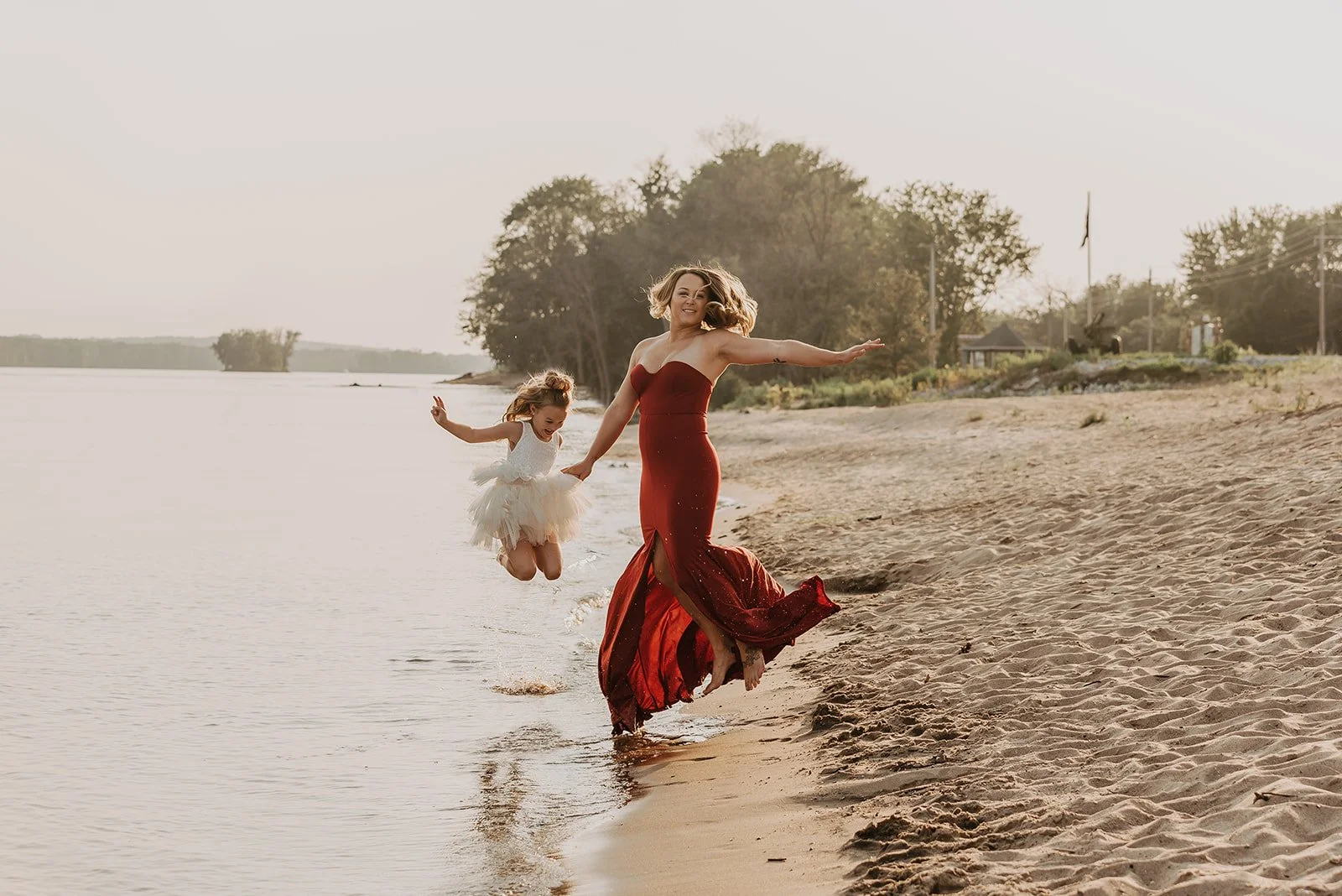 A woman in a red dress and a young girl in a white dress are jumping and playing near the water's edge on a sandy beach, with trees and houses in the background during sunset.