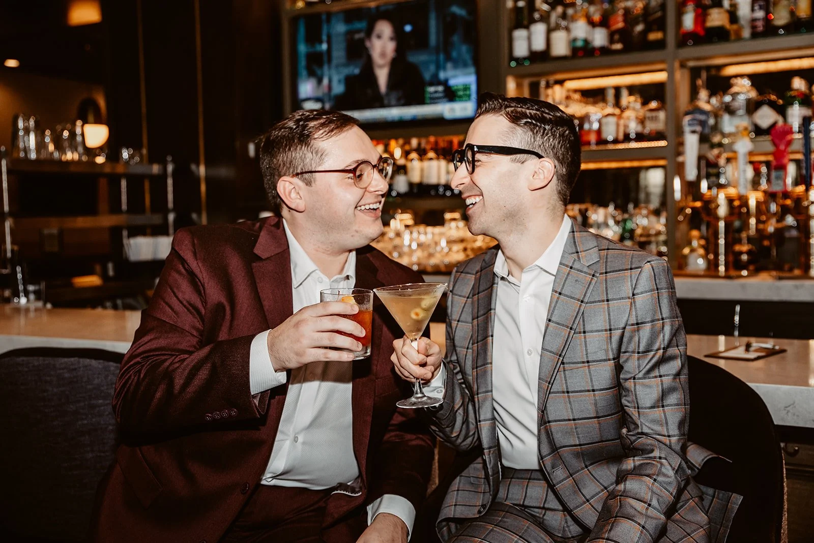 Two men in suits sharing a drink at a bar, smiling and enjoying each other's company.