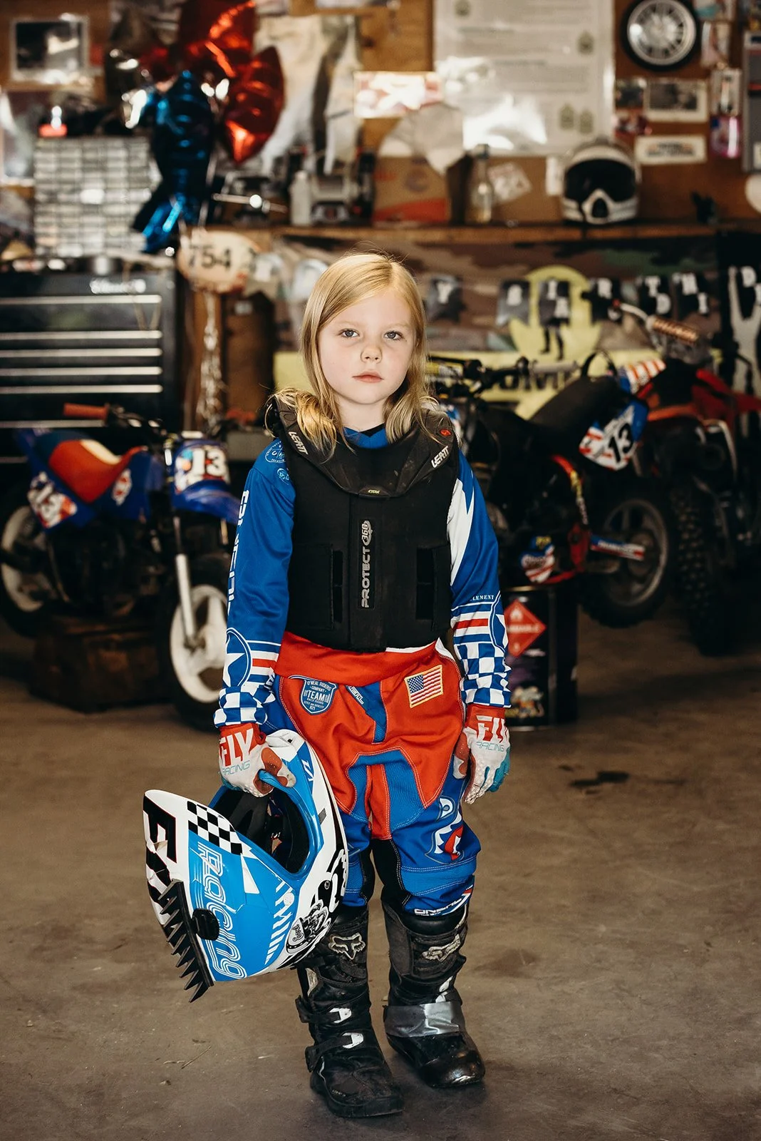A young girl dressed in motocross gear, holding a helmet, standing in a garage with dirt bikes and racing equipment in the background.