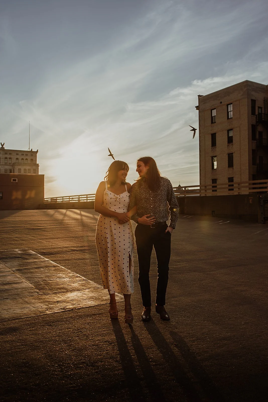 Two women walking and smiling in a parking garage during sunset, with birds flying overhead and urban buildings in the background.