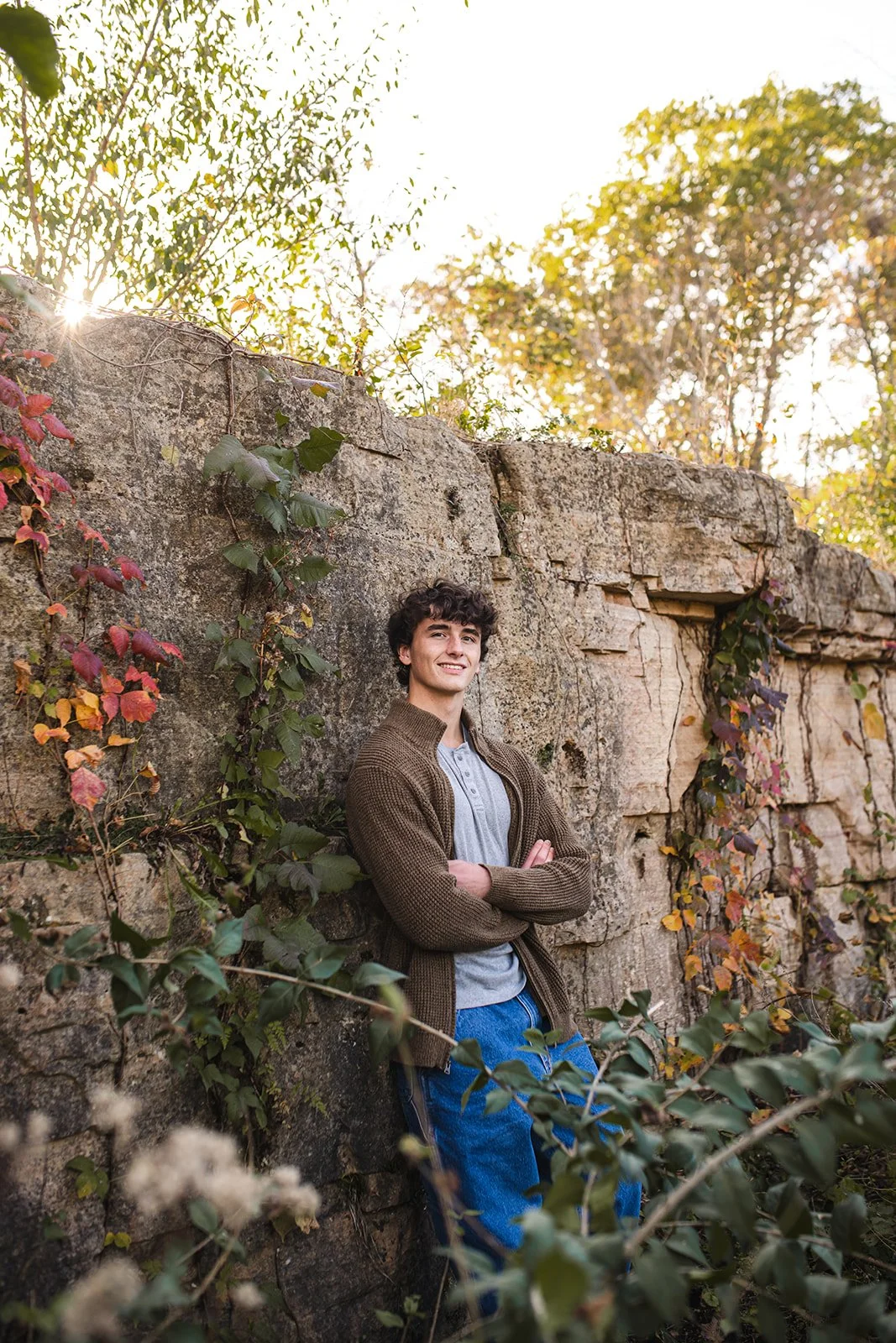 A young man with curly hair and a smile, wearing a brown jacket, gray shirt, and blue pants, standing with arms crossed against a stone wall with foliage, during autumn with sunlight filtering through trees in the background.