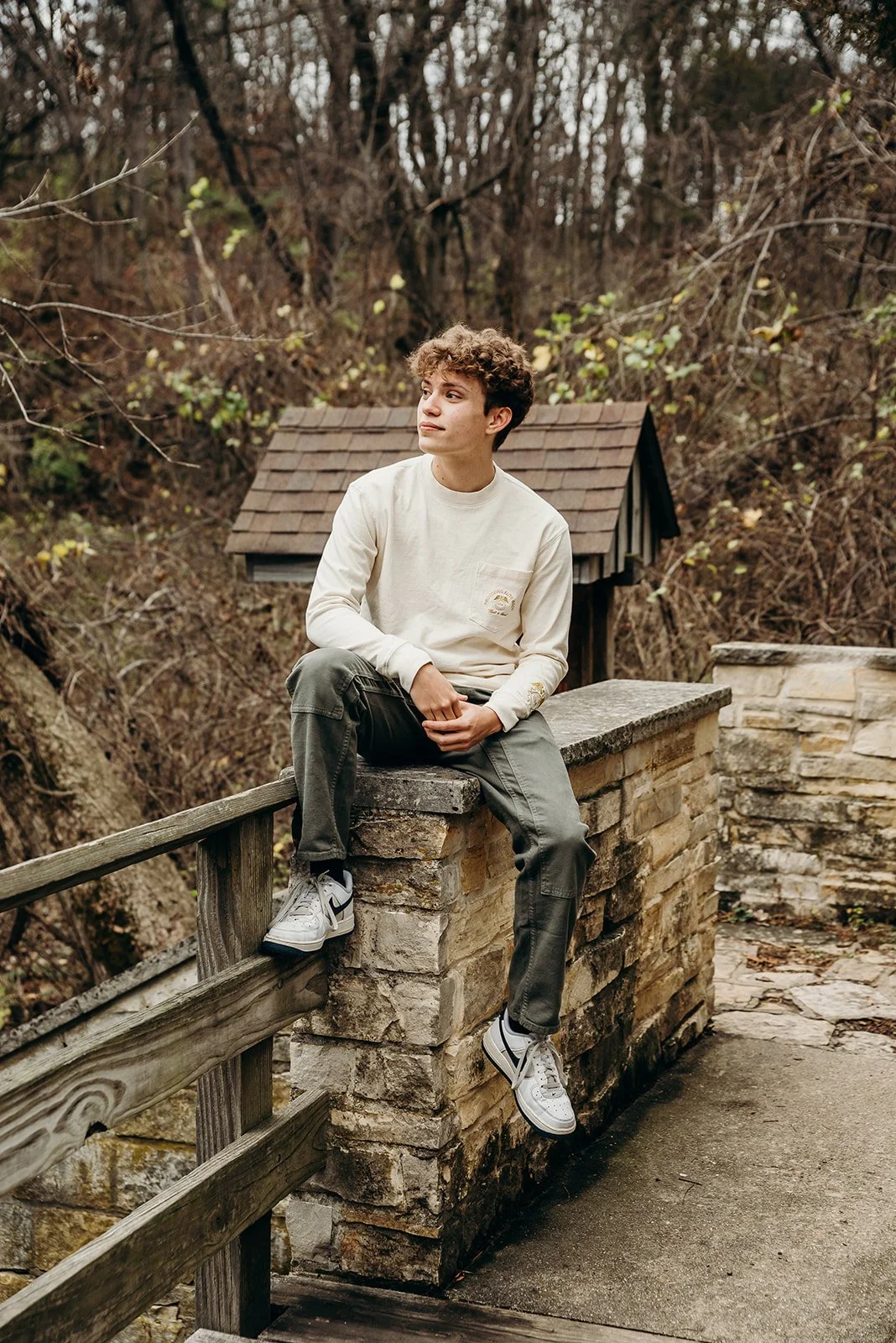 A young man sitting on a stone wall outdoors near a wooded area, wearing a long-sleeve beige shirt, dark cargo pants, and white sneakers, with a thoughtful expression.