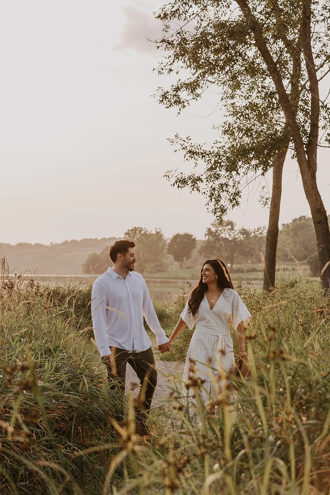 A couple holding hands and walking through a grassy field on a sunny day, smiling at each other.