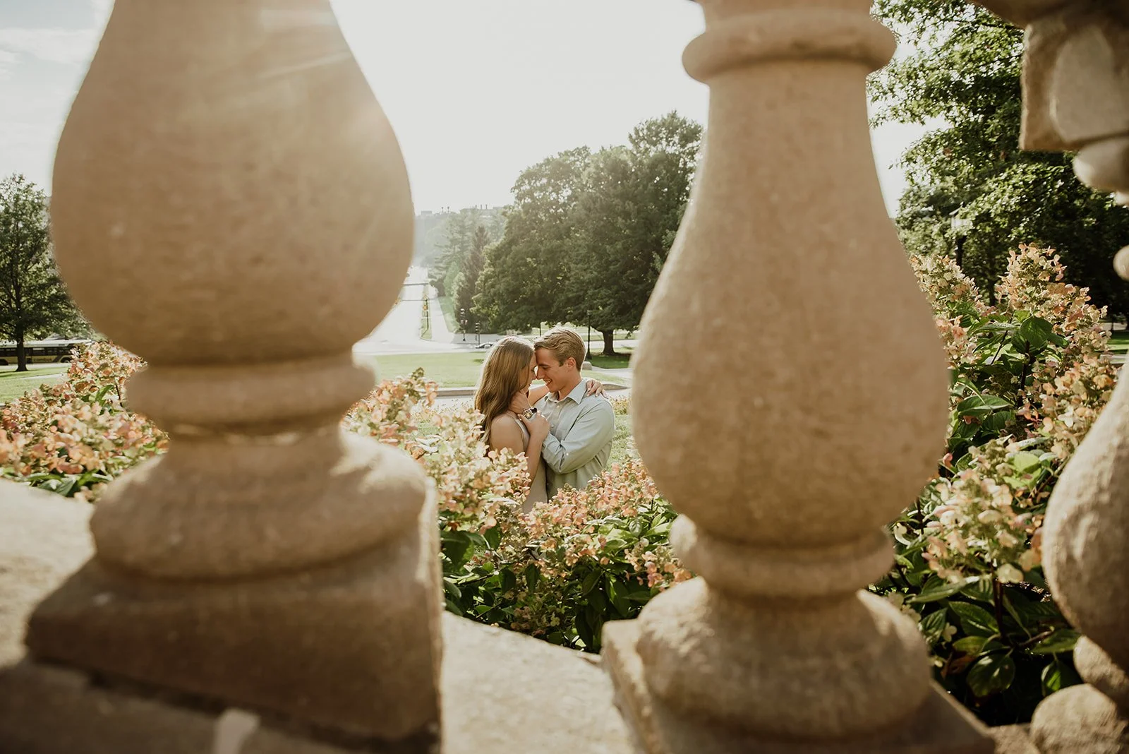 A romantic couple standing among pink flowers, seen through a stone balustrade, with a park, trees, and a pathway in the background.
