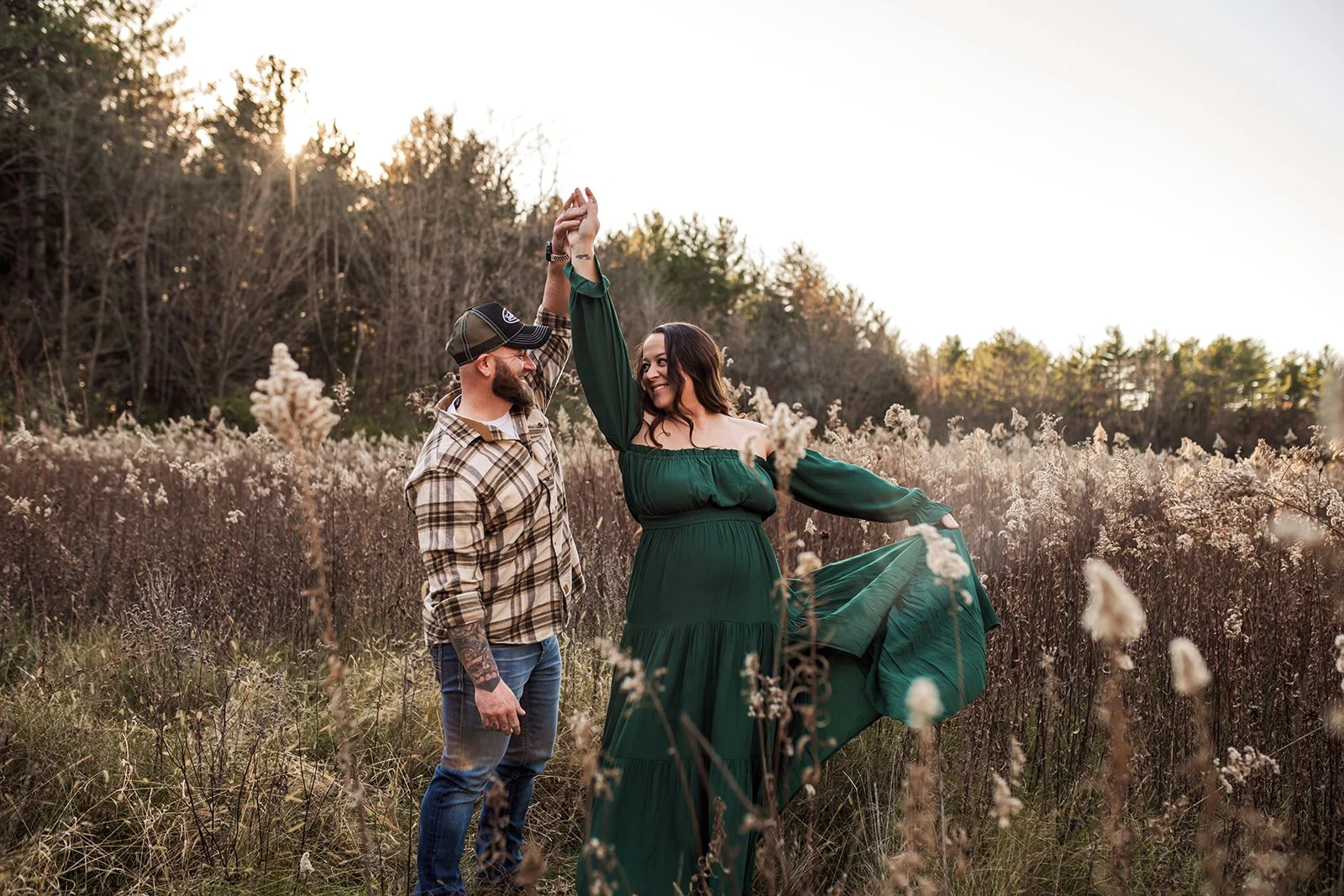 A couple dancing in a field during sunset, with the woman wearing a green dress and the man in a plaid shirt and cap, holding hands and smiling.