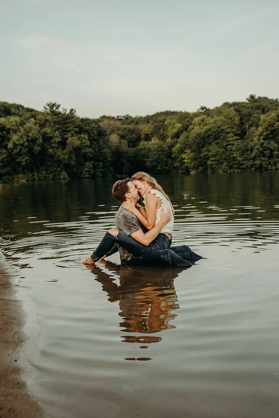 A couple hugging and smiling in a lake with lush green trees in the background during what appears to be late afternoon.