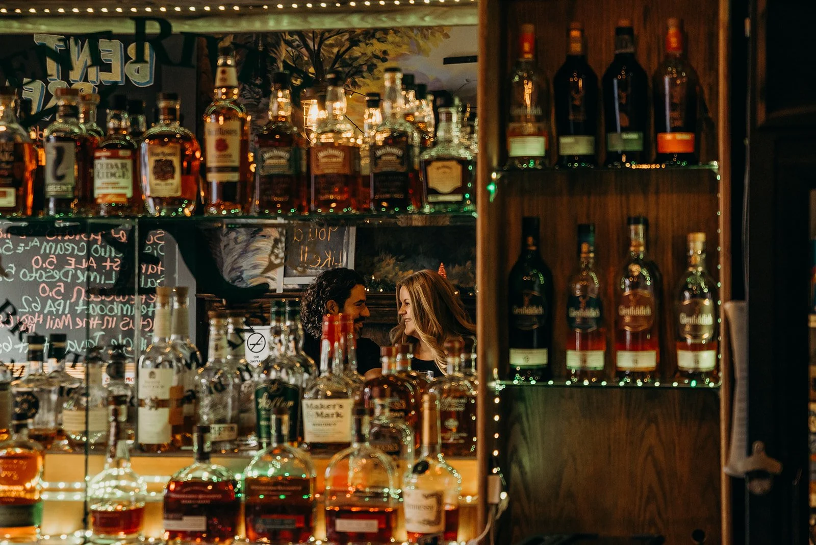 Two women inside a bar, smiling and facing each other, with shelves of whiskey bottles behind them, seen through a mirror reflecting the bar's decor and lighting.
